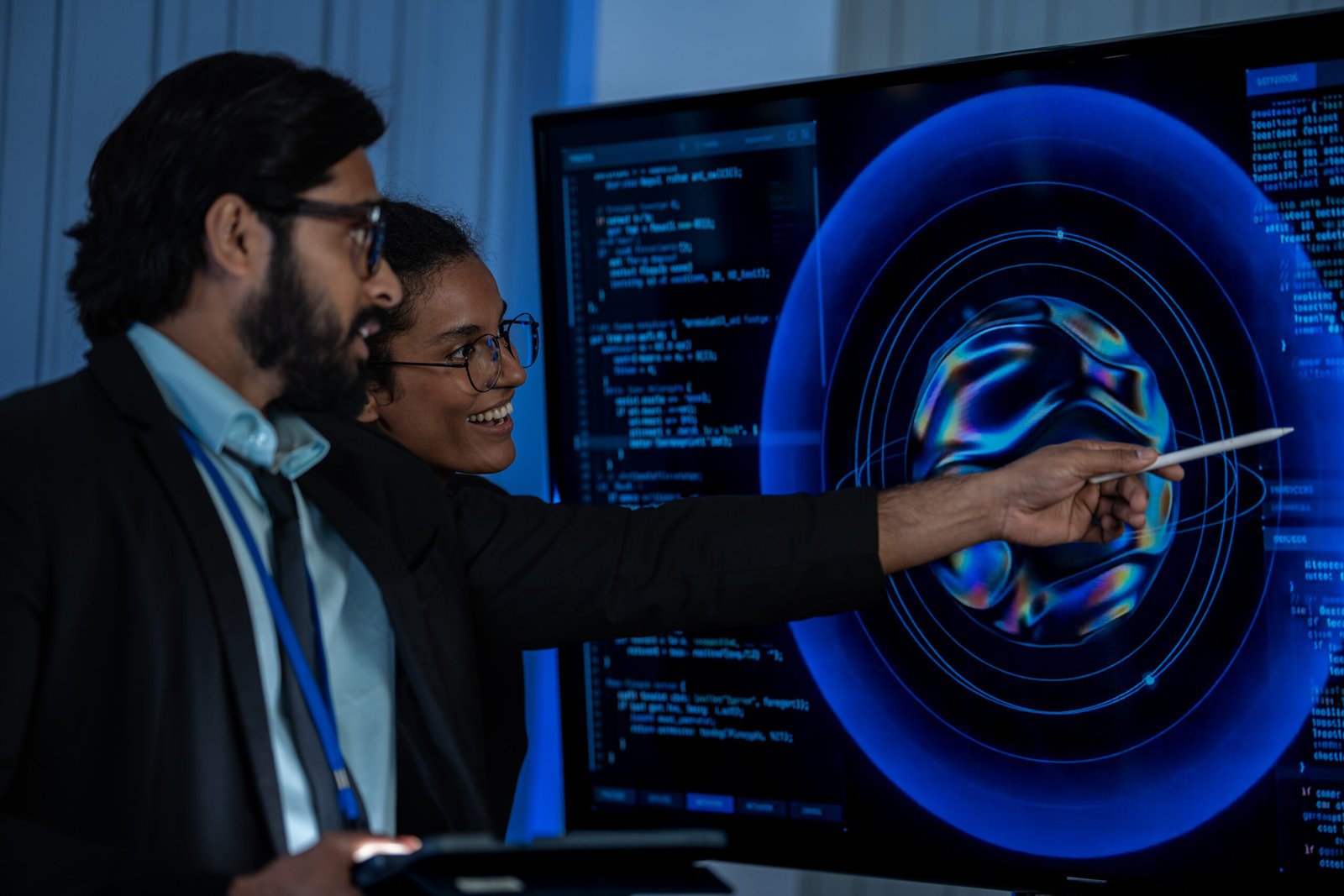 South Asian male and Black female developers review AI hologram displays on a large wall screen in a dark tech presentation room.